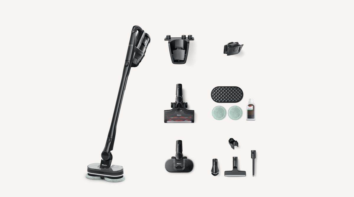 A cordless stick vacuum cleaner with multiple attachments and accessories arranged neatly on a light gray background. The vacuum is black with a small red accent, and the accessories include various floor heads, a crevice tool, and a dust cup.