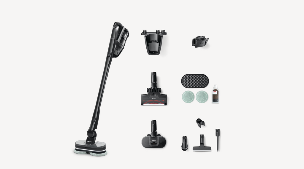 A cordless stick vacuum cleaner with multiple attachments and accessories arranged neatly on a light gray background. The vacuum is black with a small red accent, and the accessories include various floor heads, a crevice tool, and a dust cup.