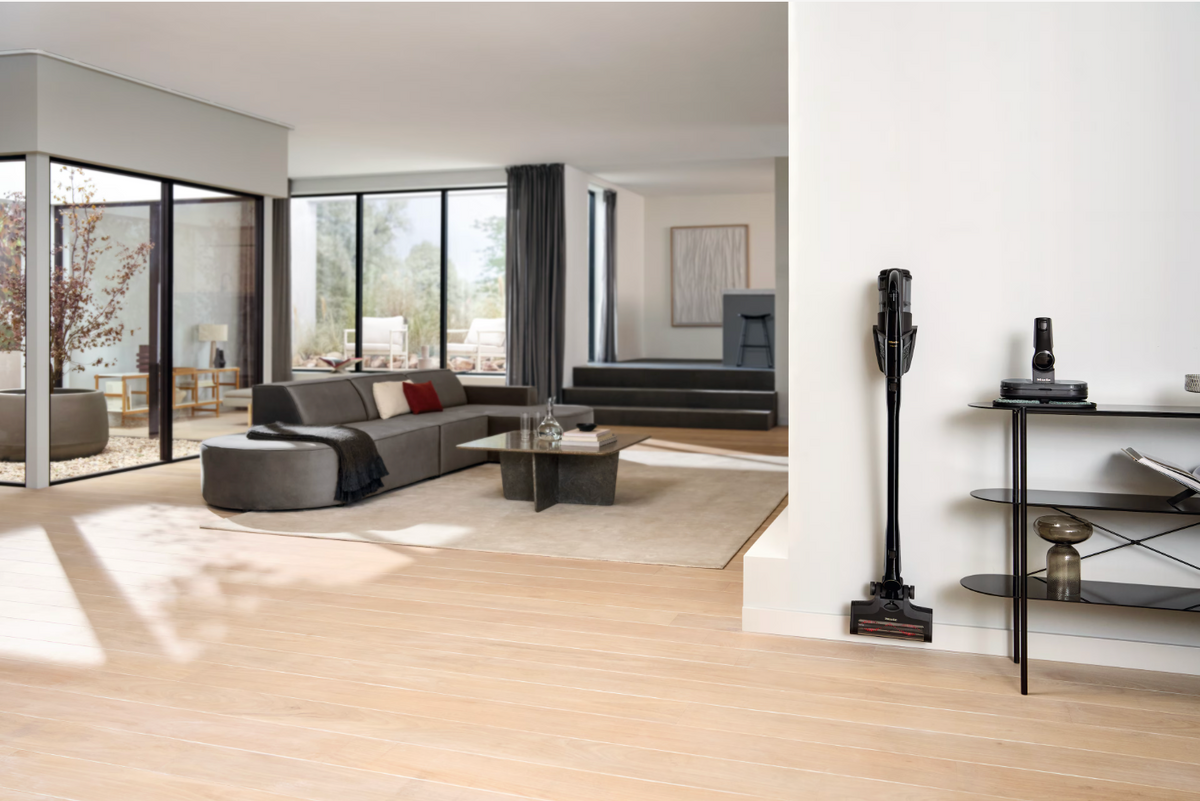 A modern open-plan living room with light oak wood flooring, featuring a gray sectional sofa, a low coffee table, and a black cordless stick vacuum cleaner standing on a shelf against a white wall.