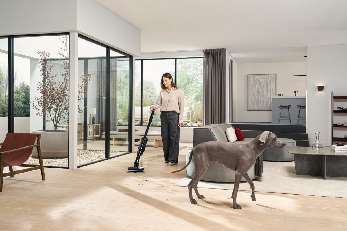 A woman using a cordless stick vacuum cleaner in a bright, modern living room with a gray dog standing nearby. The room features light wood floors, a gray sofa, and large black-framed glass windows.