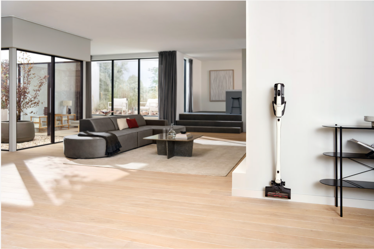 A cordless stick vacuum cleaner in white and black stands on a wooden floor in a modern, sunlit living room with a gray sectional sofa and large windows.