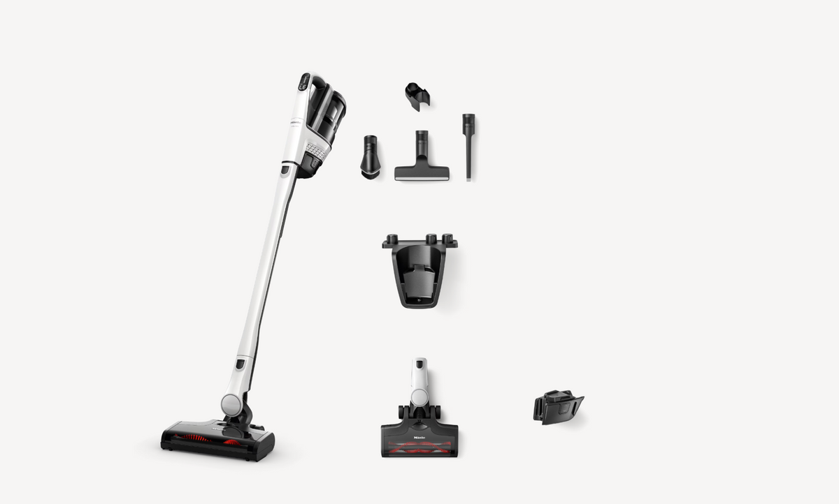 A cordless stick vacuum cleaner with multiple attachments arranged neatly on a light gray background. The vacuum features a white and black body with a silver handle, and the accessories include different floor heads, a crevice attachment, a dust bin, and a charging dock.
